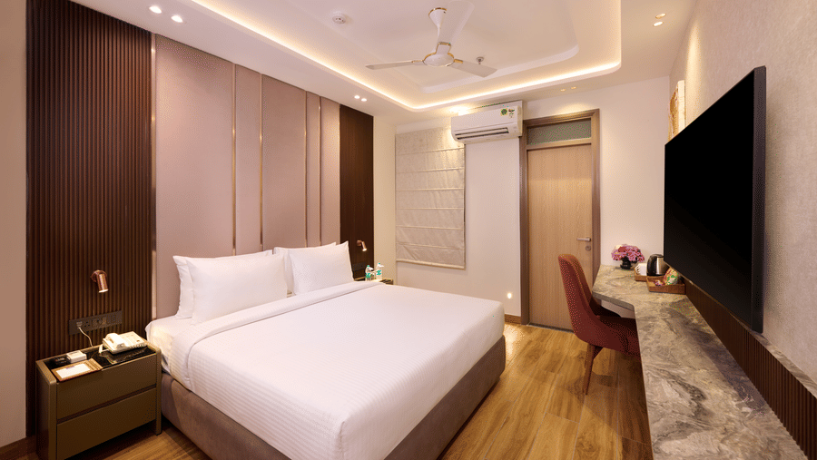 A bedroom in Tulip Inn Gurugram showing a wall-mounted flat-screen television across from a double bed with a high, padded headboard.