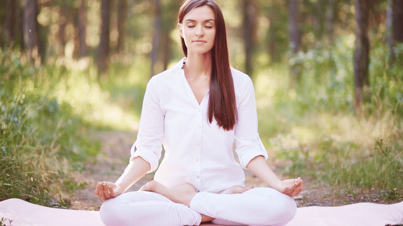 A woman meditating on a yoga mat with trees and bushes in the background