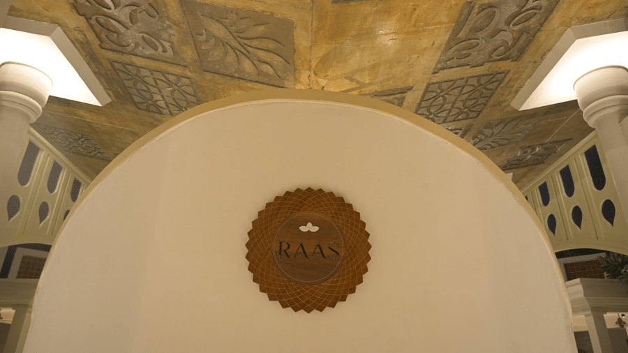A white curved wall with a circular brown emblem below a decorative ceiling at Hotel Sonar Bangla, Mayapur.