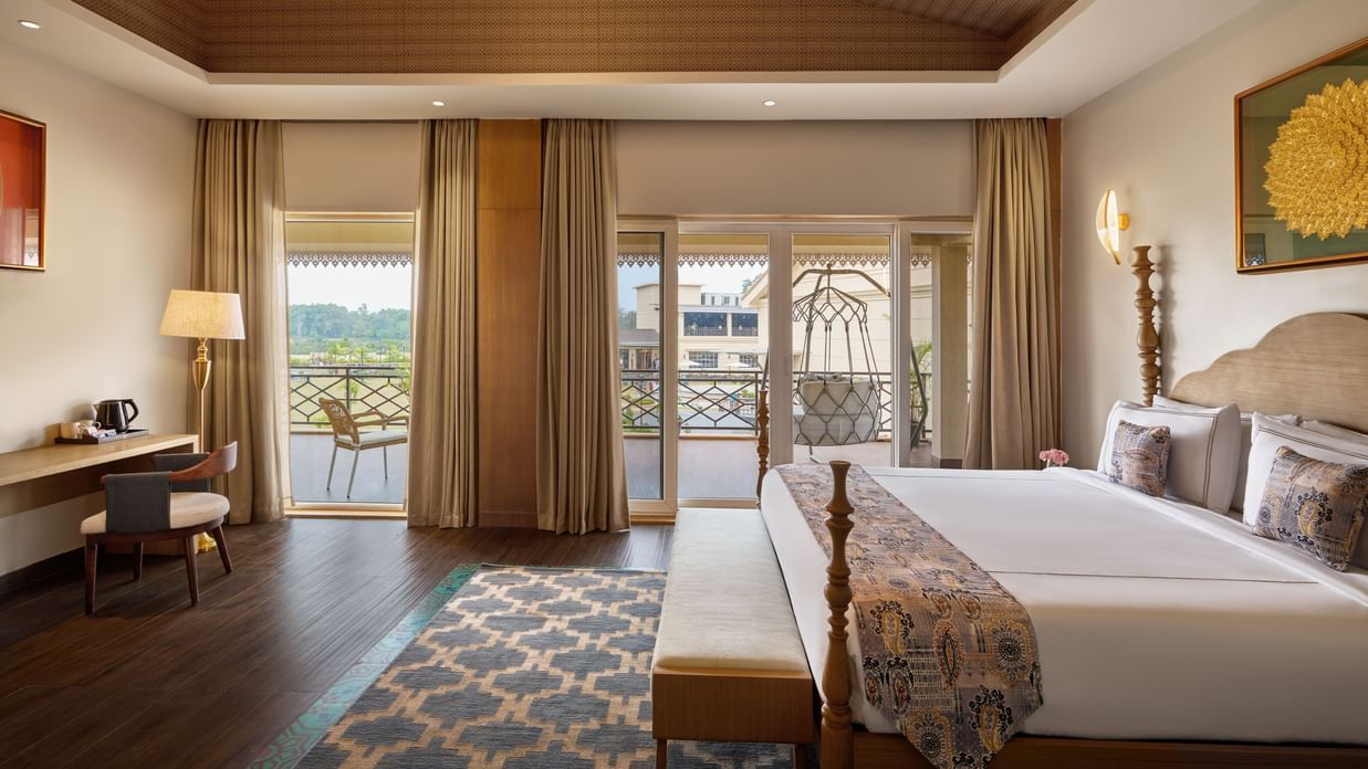 A well-lit hotel room featuring a king size bed with a patterned throw, a wooden desk and chair, and a balcony with a swing offering a view of a cityscape. The room has a wooden ceiling and a patterned rug - Voyage The Solitaire Bliss.