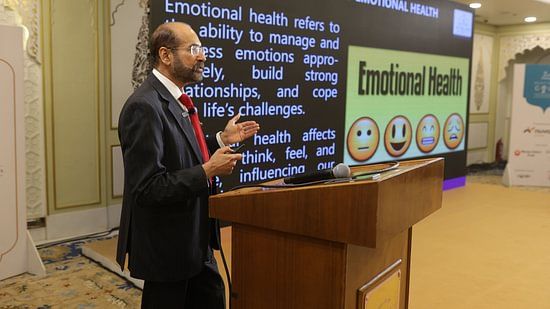 An image of a senior citizen standing in a podium and giving a speech and at the background there is some presentation going on about Emotional Health - Heritage Village Resorts & Spa