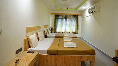 A side view of a room at Riverside Inn, featuring two beds placed beside each other, a bedside table, windows with curtains at the end, and a wall-mounted AC.