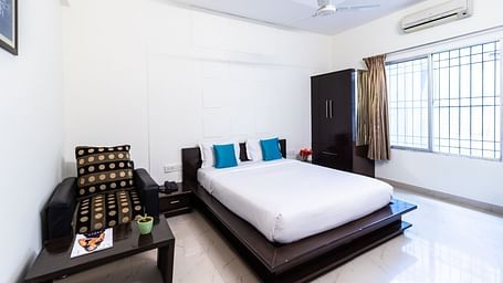 A room with a bed, an armchair, a wardrobe, and a large window at Sanctum Suites Domlur, Bangalore.