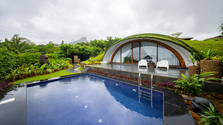 An eco-friendly luxury resort villa with a green grass-covered dome roof facing a private blue swimming pool under an overcast sky.