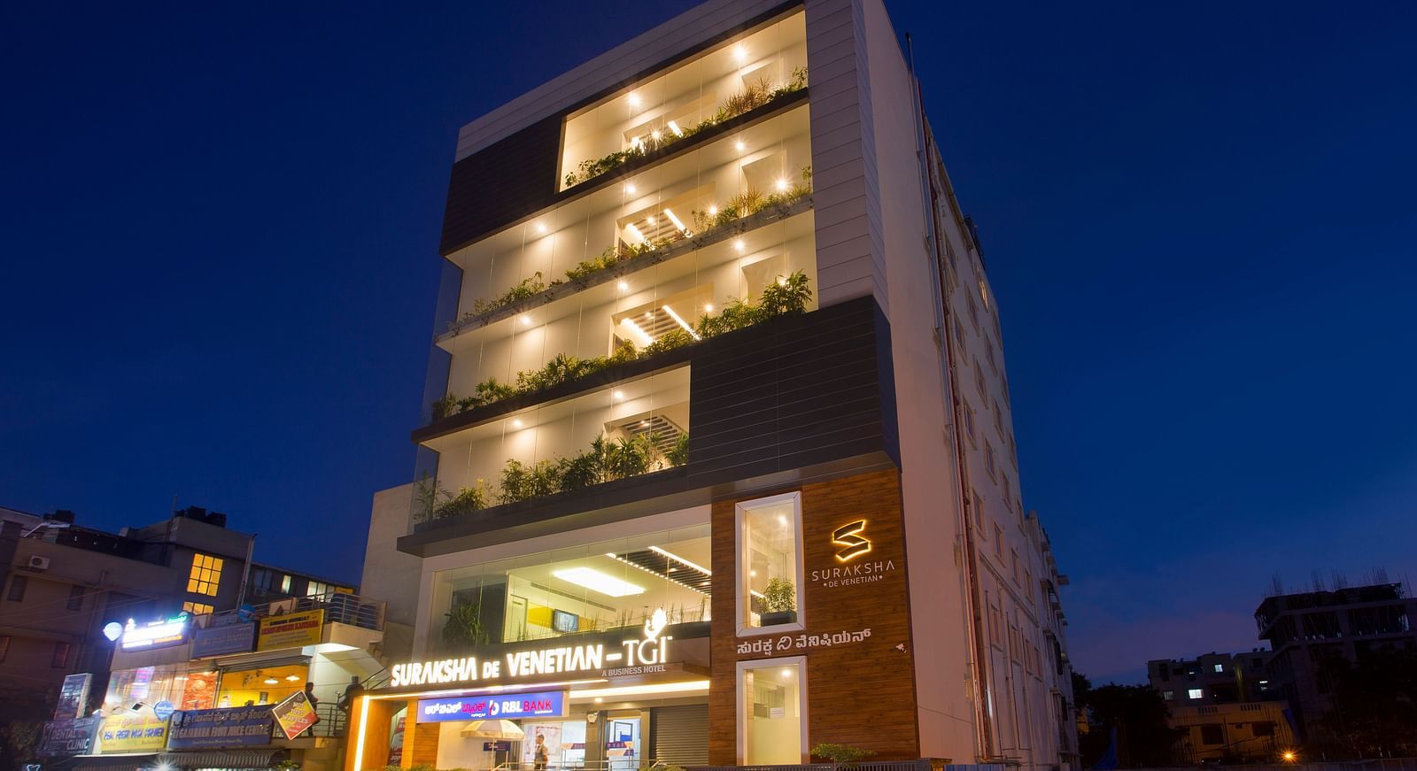 An exterior view of a modern multi-story building illuminated at dusk with signage and street activity - De Venetian by TGI, Brookefield