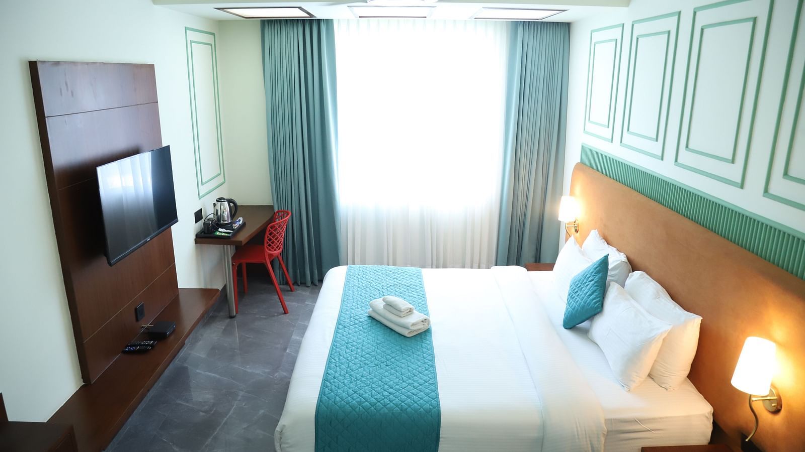 A bright room featuring a double bed with a blue runner, bedside tables, and a television - Asian Suites IFFCO Metro, Gurgaon