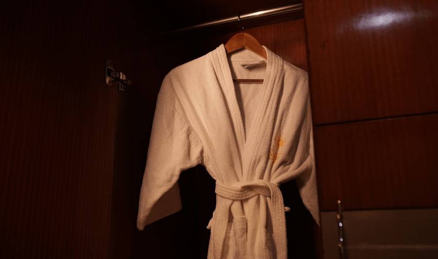 A white bathrobe on a wooden hanger inside a dimly lit wardrobe, conveying a sense of luxury and relaxation. The wardrobes wood finish adds warmth.