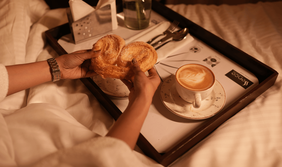 A tray on a bed with a person holding a heart-shaped pastry, next to a cappuccino. A calm, cosy breakfast setting with soft, warm lighting.
