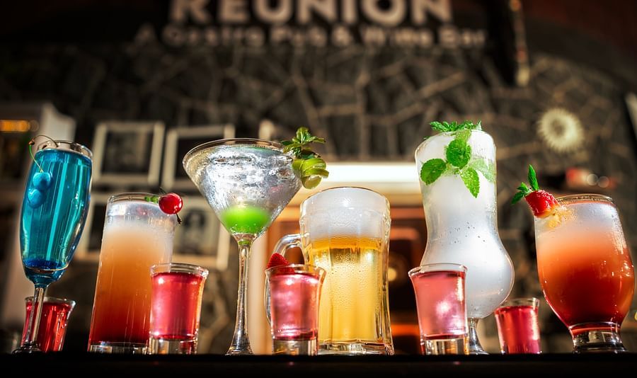 A vibrant display of colourful cocktails and drinks, garnished with cherries, lime, mint, and strawberries, set against a blurred pub background with the sign REUNION visible at Uday Samudra Leisure Beach Hotel.