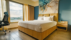 A well-lit hotel room with a large bed, wooden flooring, a work desk, a window with a view, and a large abstract art panel above the headboard | Pride Elite Chhatrapati Sambhajinagar