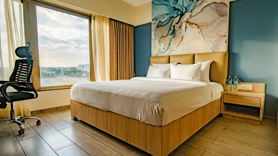 A well-lit hotel room with a large bed, wooden flooring, a work desk, a window with a view, and a large abstract art panel above the headboard | Pride Elite Chhatrapati Sambhajinagar