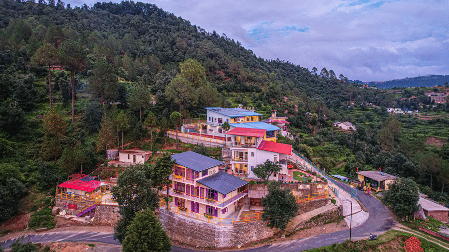 A view of buildings on a hillside with trees and a road passing through the area at Adrushya Estates, Mukteshwar.