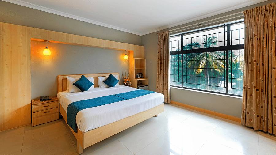 An image of a hotel room in Bengaluru featuring a queen size bed, a night stand, and sunlight from the expansive windows - Cross Roads Inn, Bengaluru