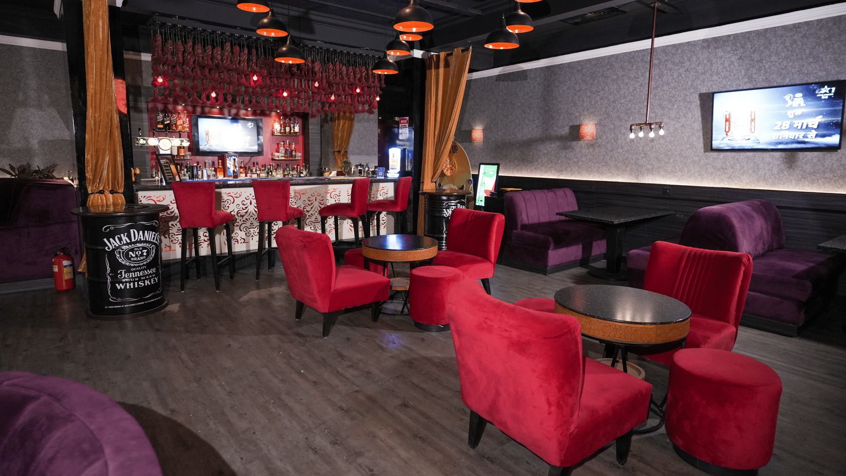 Wide view of a lounge at Park Plaza Ludhiana featuring red sofas and chairs dark ceiling with orange pendant lights a bar counter in the background and a wall-mounted TV.