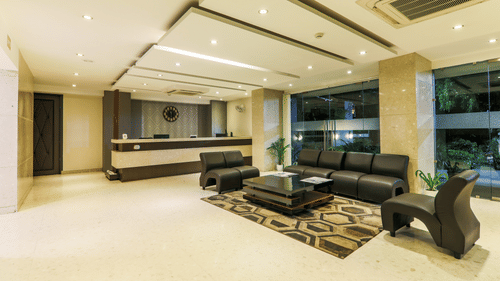 Lobby area of Magnus Star Residency, Koregaon Park Annexe, featuring a modern reception desk, black leather seating, glass walls with views of greenery, geometric rug, and warm recessed ceiling lighting.