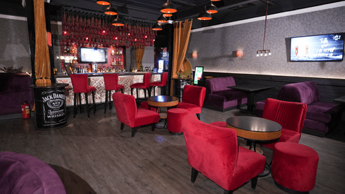 Wide view of a lounge at Park Plaza Ludhiana featuring red sofas and chairs dark ceiling with orange pendant lights a bar counter in the background and a wall-mounted TV.