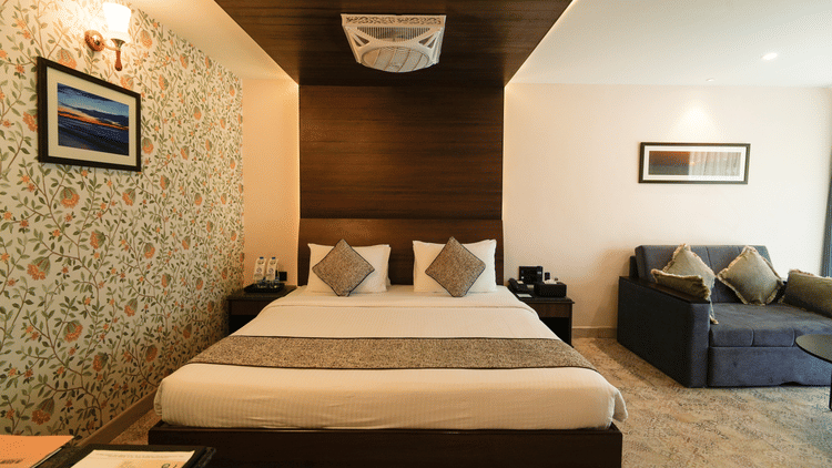 A neatly arranged bed with pillows and a comforter in a hotel room with modern interiors, a bedside table, a sofa on the side, and a photo frame on the wall