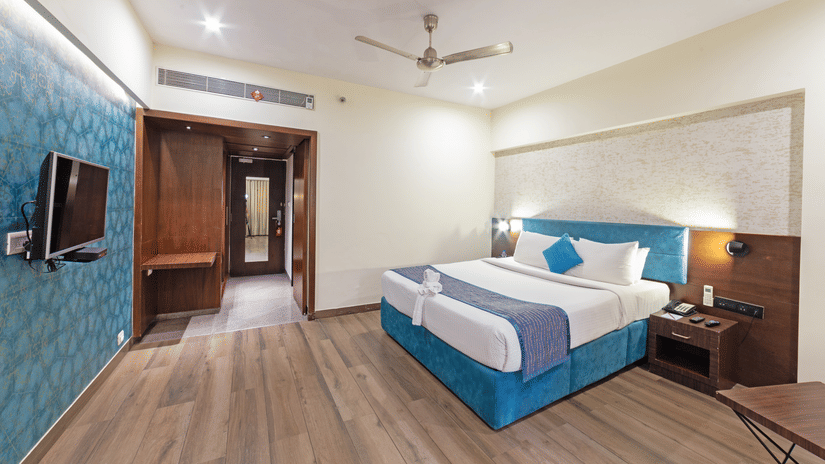 A corner spacious and well-lit hotel room with a bed, a flat-screen TV, and a seating area. The room features a wooden floor and blue accents on the walls.