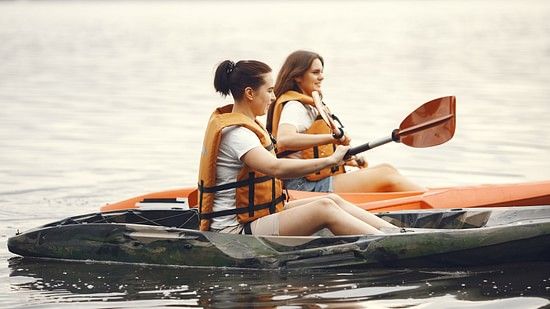 Two women in life jackets kayaking on a calm lake, surrounded by serene water and lush greenery.