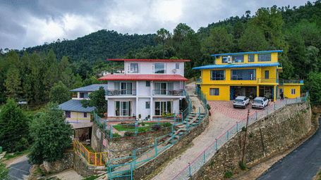 A hillside property with buildings, road, and trees surrounded by green hills at Adrushya Estates, Mukteshwar.