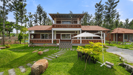 Villa exterior in daylight with a wide lawn, a stone pathway, scattered rocks, tall trees and a wooden and brick building in the background.