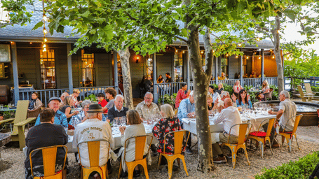 A crowd seated at outdoor dining arrangement and eating under tree shade at Tallman Hotel