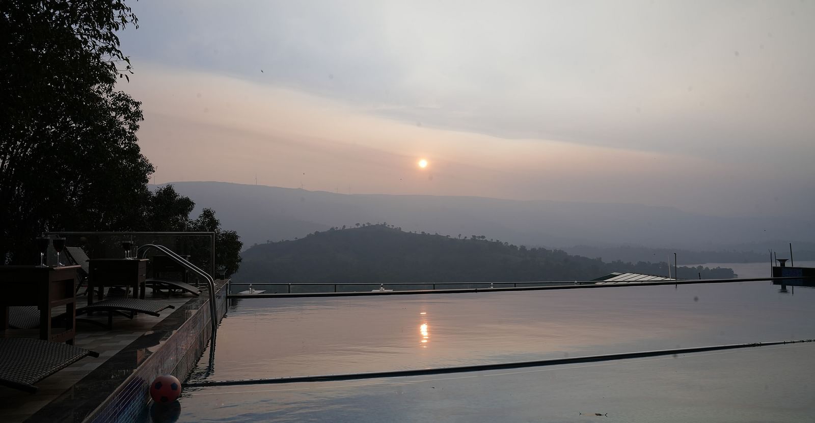 An image of the infinity waterfall swimming pool with the backdrop of the mountains and the setting sun - Nisarga Lake Resort