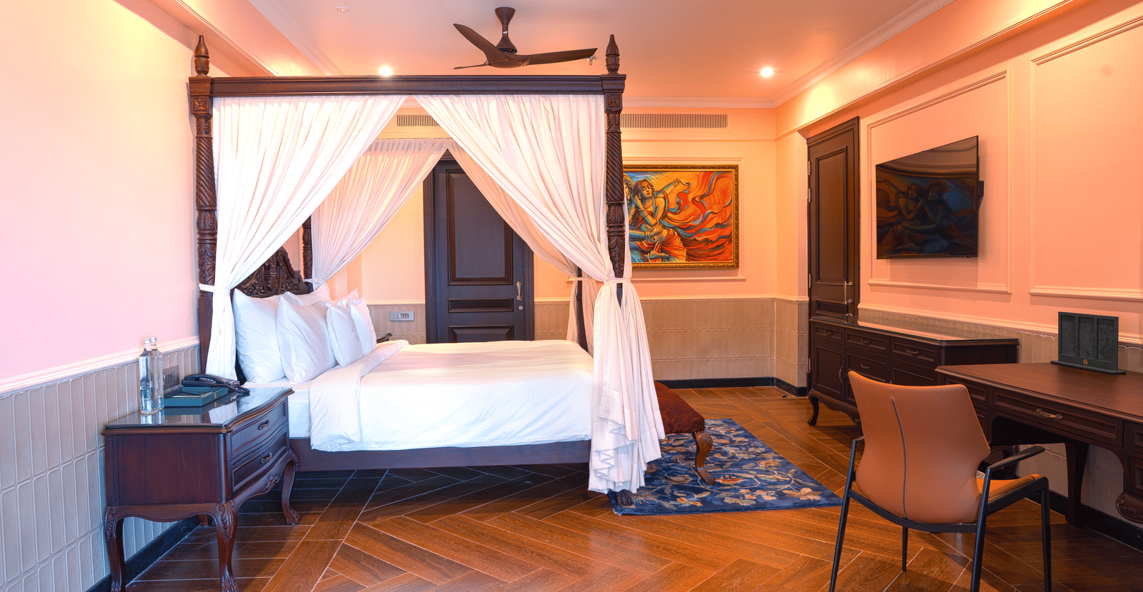 A luxurious 4-poster king size bed with white canopy drapes in a room with wooden floors and contemporary artwork at at MAYFAIR Bay Resort, Paradeep.