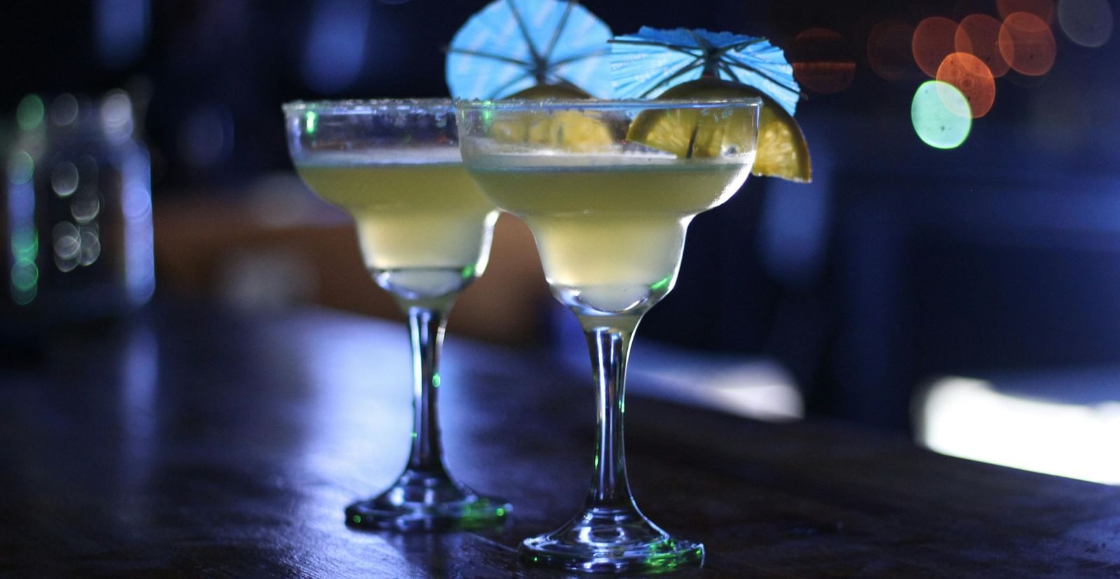 Two margaritas in stemmed glasses with lime wedges and paper umbrellas, on a dark bar counter.