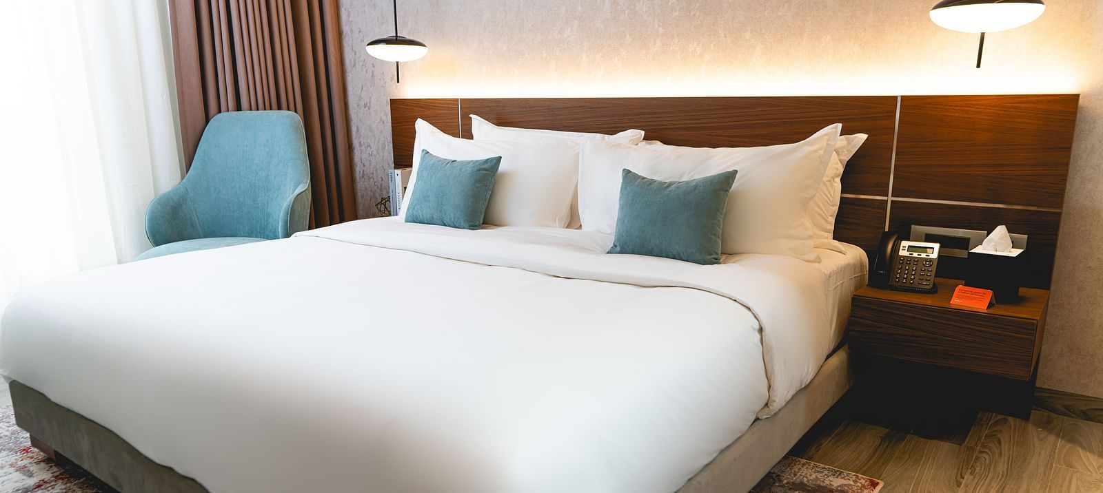 A bed and accent pillows at KertelSuites, featuring a wooden headboard, two bedside tables with lamps, and a armchair by the window