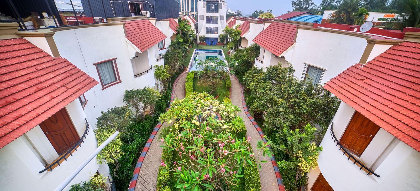 Elevated view showcasing a row of villa-style accommodations with striking red tiled roofs and lush green pathways between them at Ilara Hotels & Spa, OMR, Chennai.