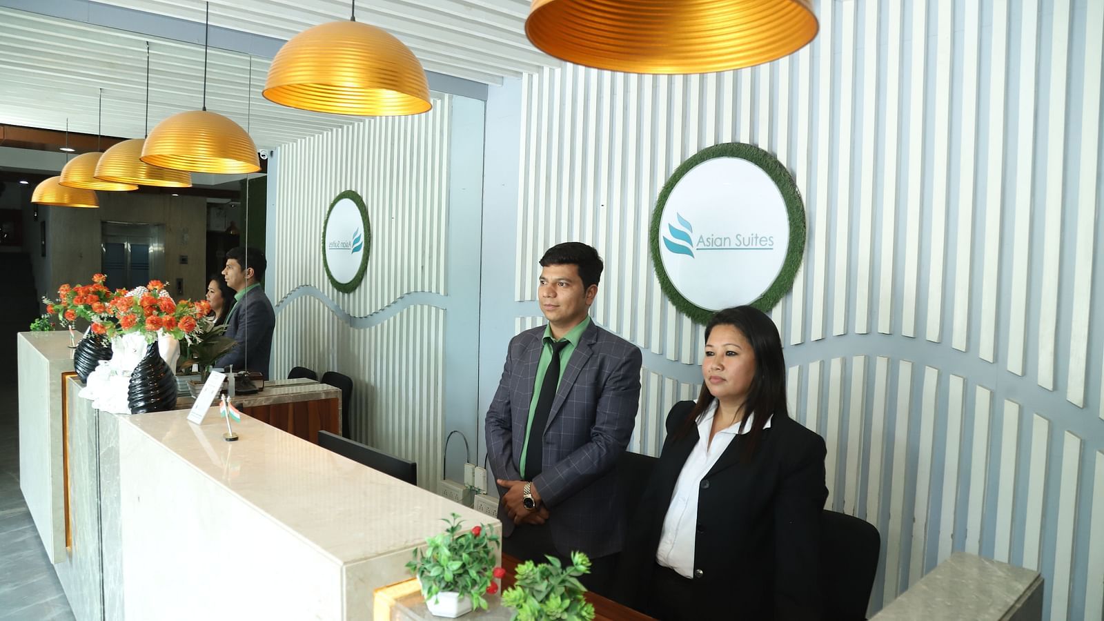Two individuals stand behind a reception desk in a well-lit area with decorative lighting - Asian Suites IFFCO Metro, Gurgaon
