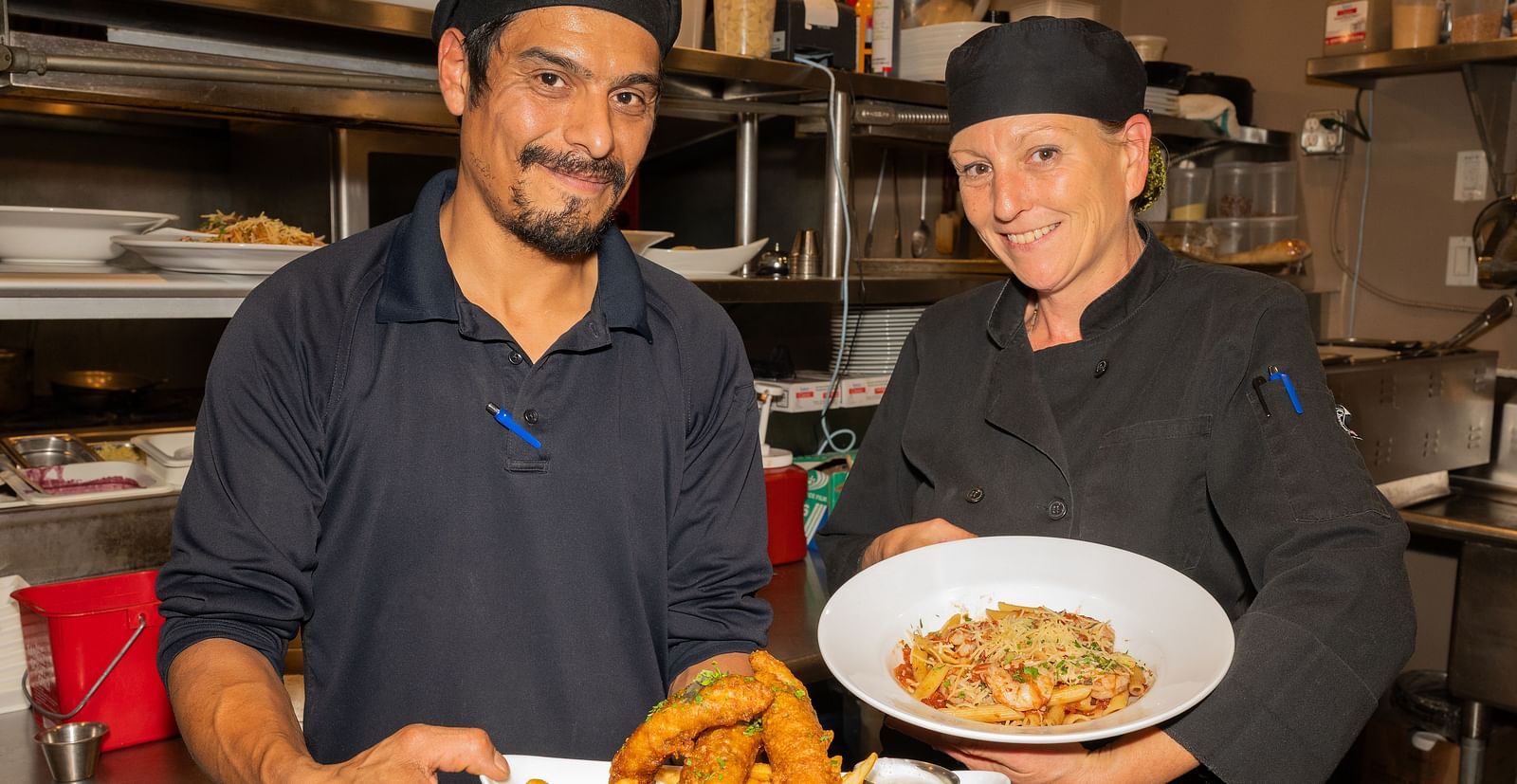 Two chefs smiling while holding plated dishes in the kitchen at The Groveland Hotel.