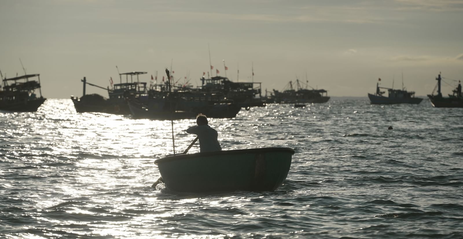 Fisherman rowing a small boat on the sea with several docked boats in the background.