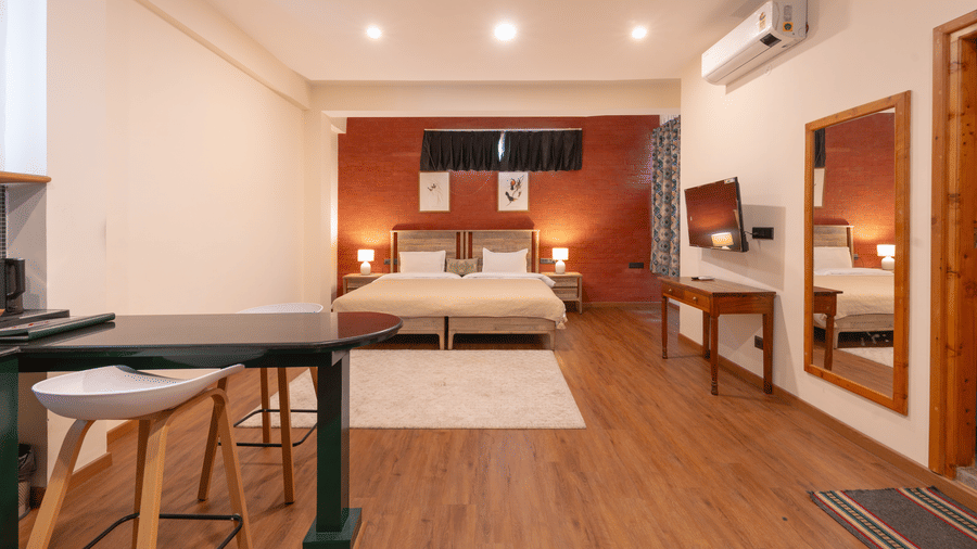 A wide-angle view of a studio room featuring wooden flooring, a small kitchenette, and twin beds against an orange accent wall - Johnson Lodge & Spa, Manali