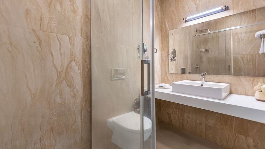 A bathroom with beige and brown textured walls, a glass shower stall, a white rectangular sink, and a mirror - Mango Hill Resort, Coimbatore