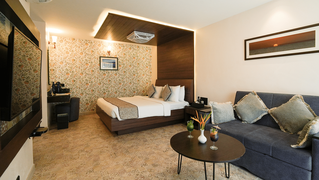 A room with a bed, a sofa with cushions, a table with a flower vase, and a TV mounted on the wall