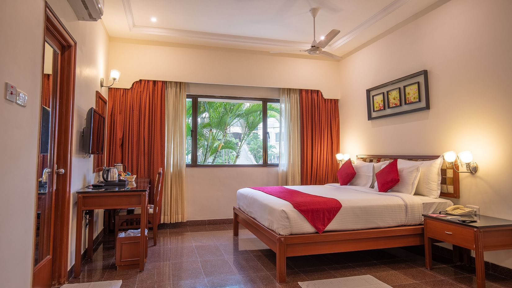 A well-lit Executive Suite features a king sized bed, bedside tables with lamps, a window with curtains, a ceiling fan, a wall-mounted TV, and a small desk with a chair