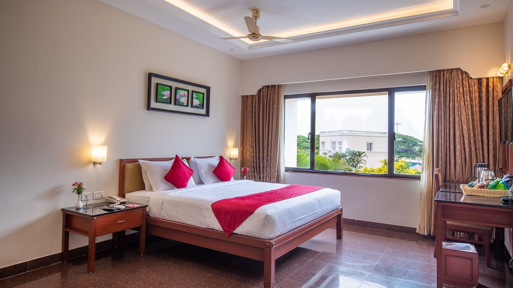 A bright Executive Room with a king sized bed, pillows, bedside tables with lamps, a desk with a chair, curtains framing a large window, wall art, and a ceiling fan