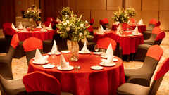 A banquet hall setup at KPG Sarovar Portico, Raj Nagar Extension, Ghaziabad featuring round dining tables, chairs, flower vases, and tableware.  