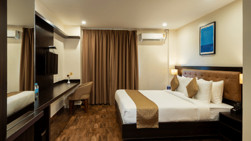 An elegant bedroom at VITS Select, Bengaluru, featuring a plush bed, rich wall paneling and curtains, a reflective mirrored surface, and a mounted air-conditioning unit for the perfect stay in Bengaluru.