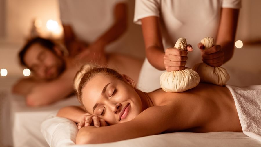 A couple undergoing Ayurvedic kizhi massage, laying on white beds, assisted by two practitioners. 