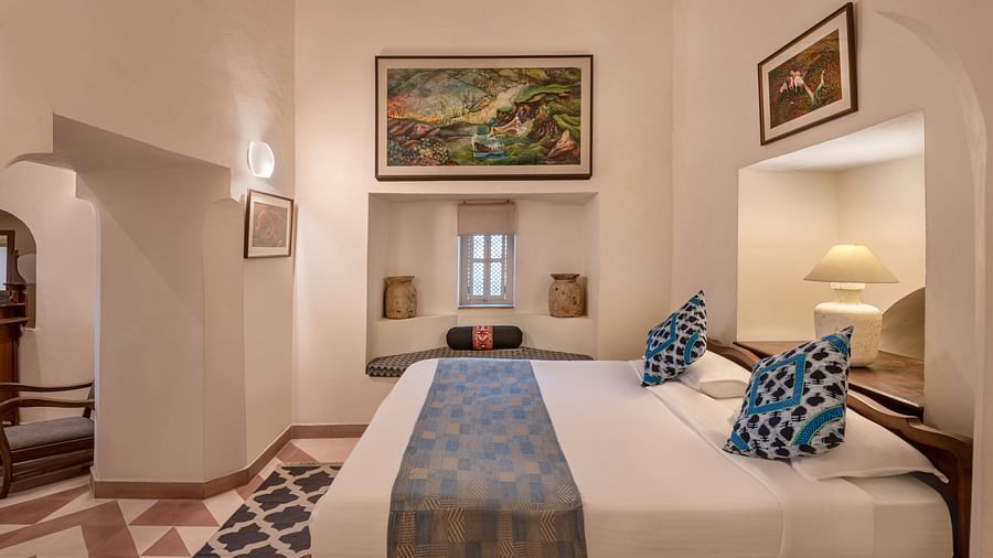 A room with a bed, two framed artworks, a window seat, and a small window in Tijara Fort-Palace - 19th Century, Alwar.