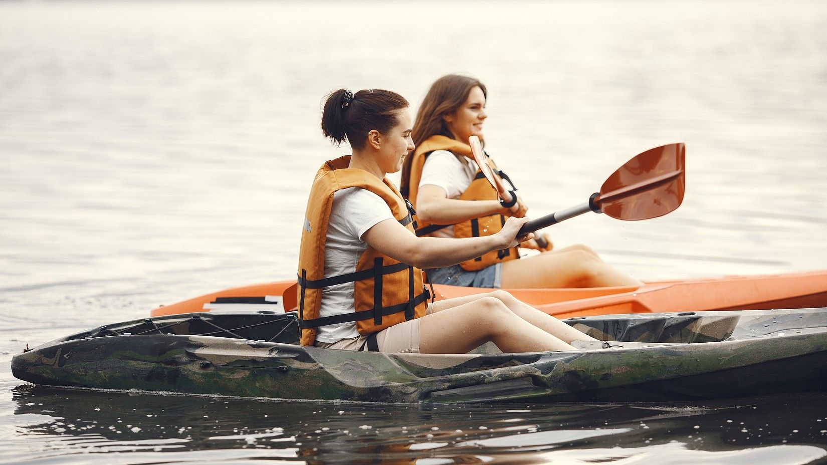 Two women in life jackets kayaking on a calm lake, surrounded by serene water and lush greenery.