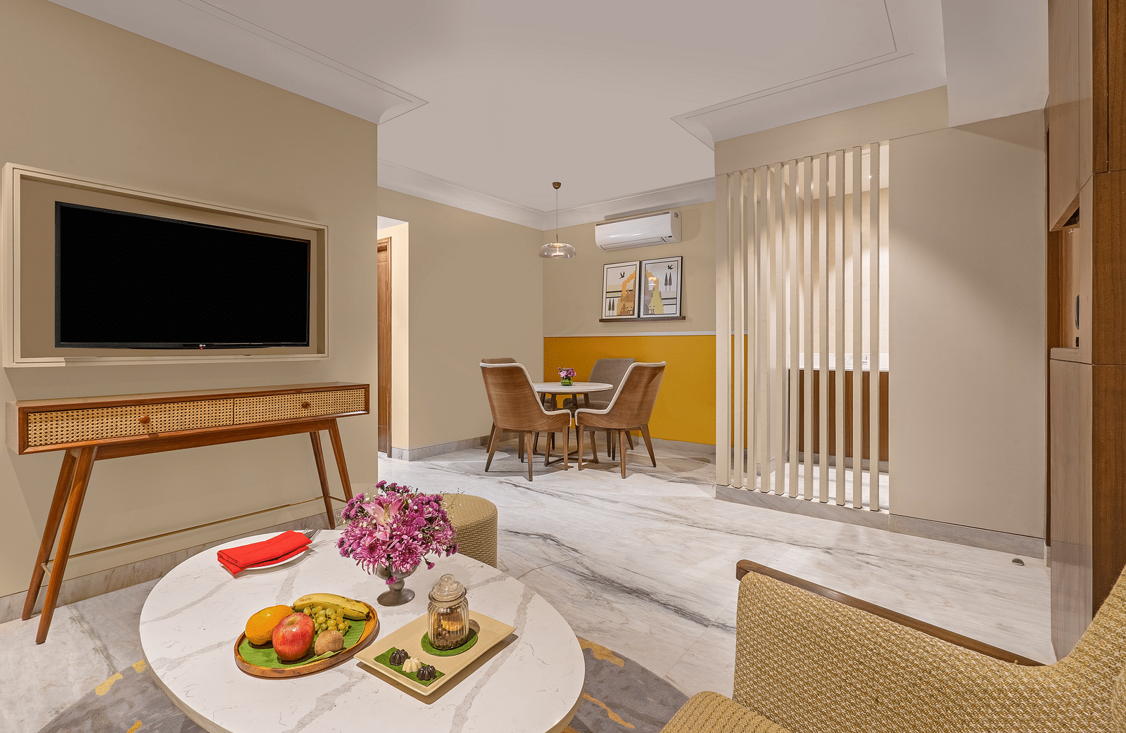 An image of a living room featuring a comfortable sofa, a coffee table with fruit and snacks, a television mounted on the wall, and a dining table visible in the background. A wooden partition separates the living area from the dining area.