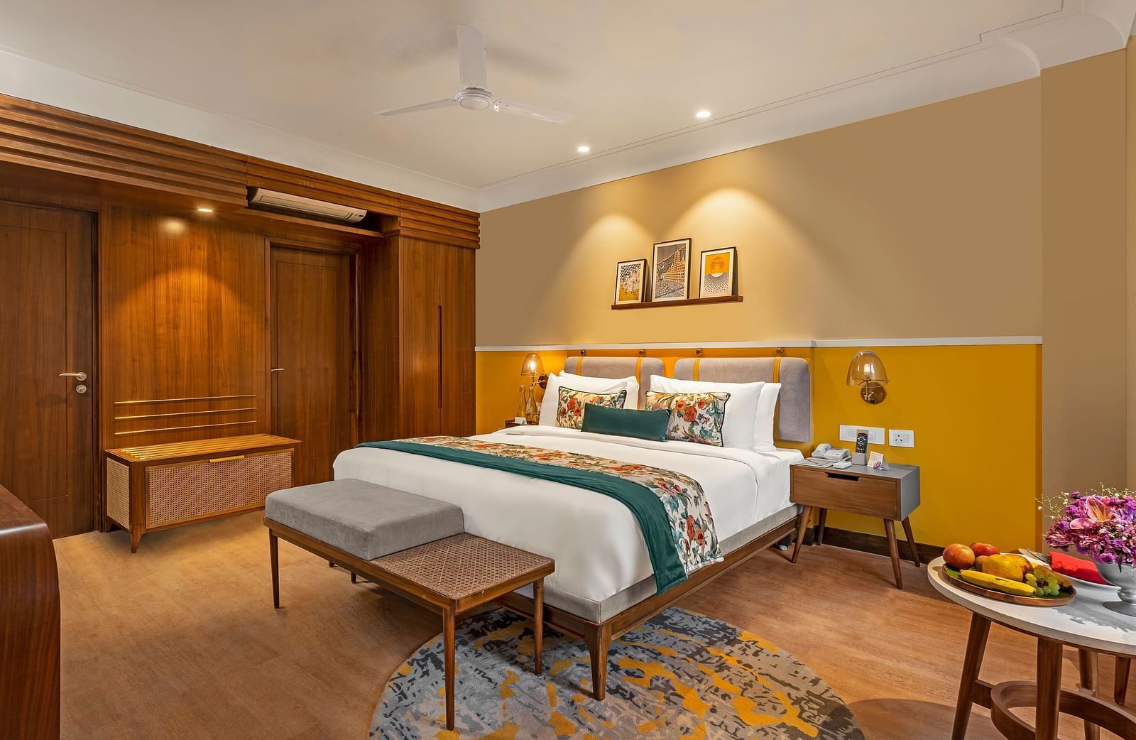 An image of a room featuring a king-size bed with a headboard and comfortable bedding. A bench sits at the foot of the bed, and a small table with a fruit platter is placed nearby.