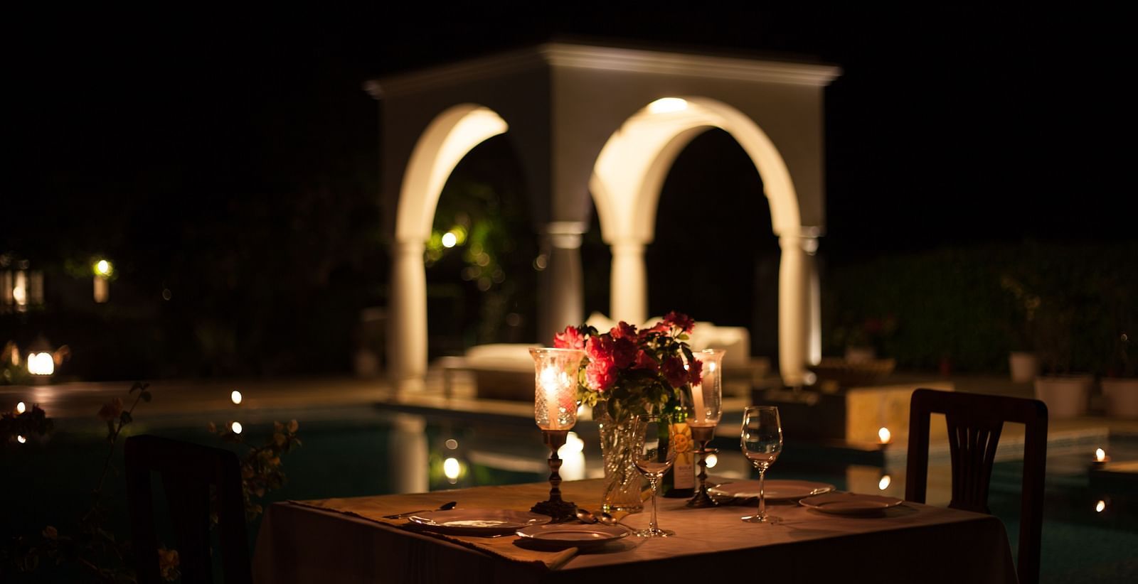 A dimly lit outdoor dining setup with a table set for two, featuring a flower arrangement and glassware, with a structure featuring arches in the blurred background- Shahpura Bagh.