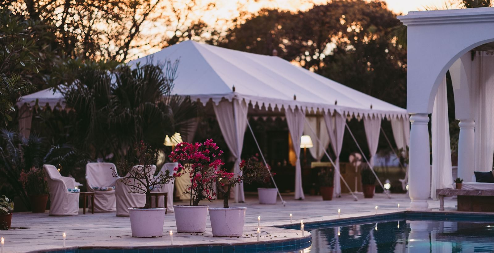 A white tent with draped sides is set up beside a swimming pool, illuminated at duskA white tent with draped sides is set up beside a swimming pool, illuminated at dusk- Shahpura Bagh.