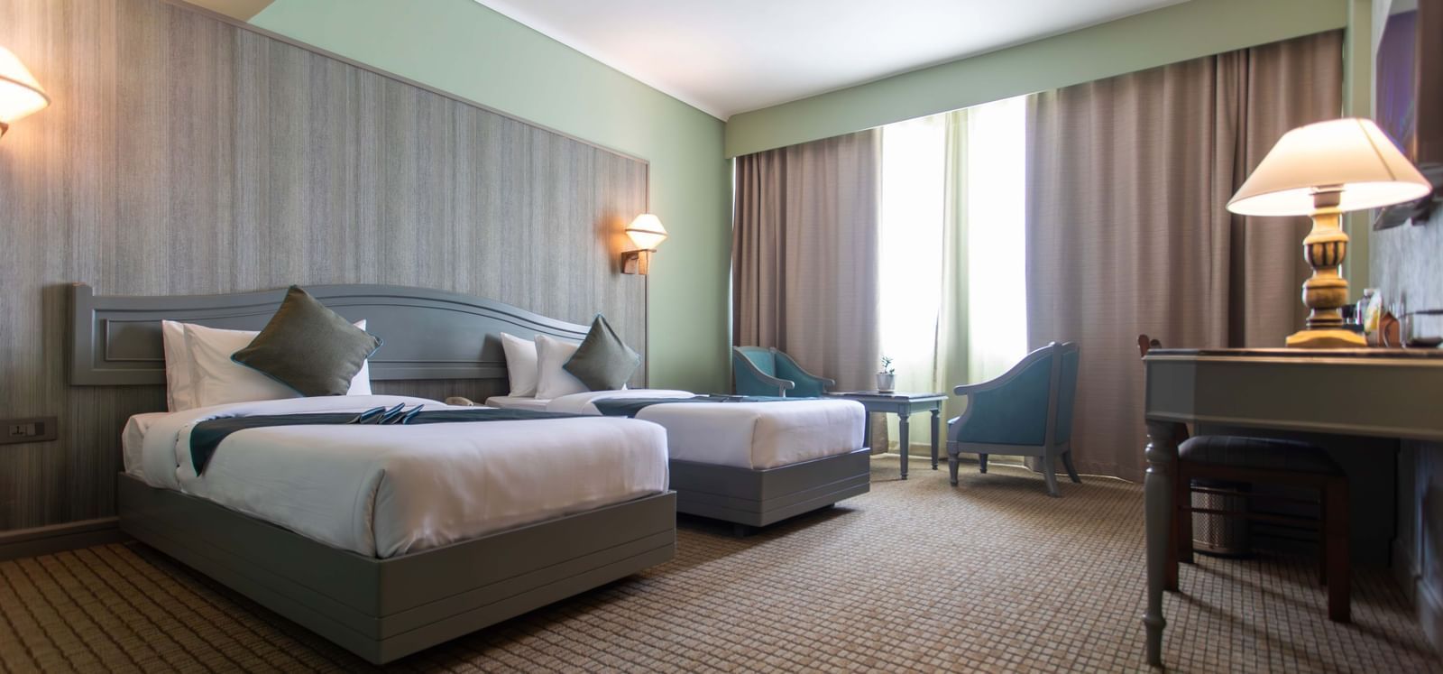 A hotel room at Shilton International, Kochi, with two beds, a sitting area, and ample natural light.