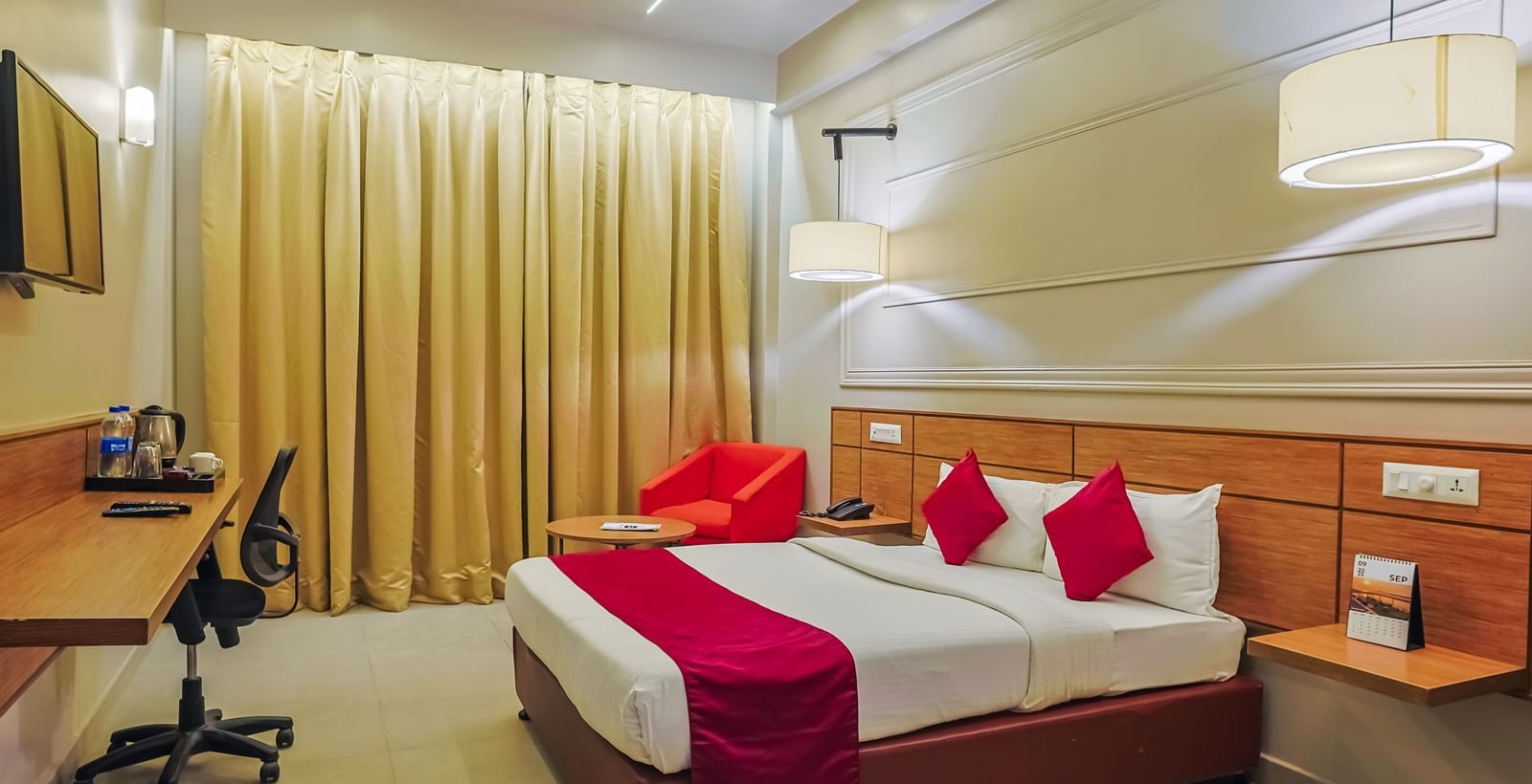 Bedroom with double bed, red runner, pillows, desk and curtains at Max Hotel Prayagraj.